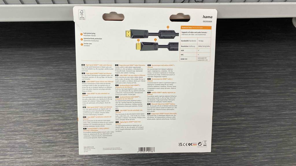 Eine weiße Verpackung mit Fokus auf ihrer Rückseitige Beschreibung eines Hama-HDMI-Kabels in 10 Meter Länge inklusive goldener Stecker steht zum Test bereit an ein Verkaufsregal gelehnt auf einem grauen Boden.