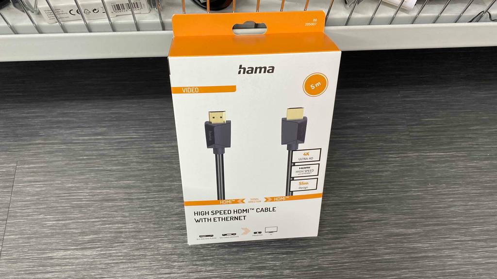 5 Meter langes Hama-HDMI-Kabel im Test: Eine weiße Verpackung mit orangen Design-Elementen lehnt mit der Vorderansicht inklusive goldener Stecker auf einem grauen Untergrund vor einem Verkaufsstand.