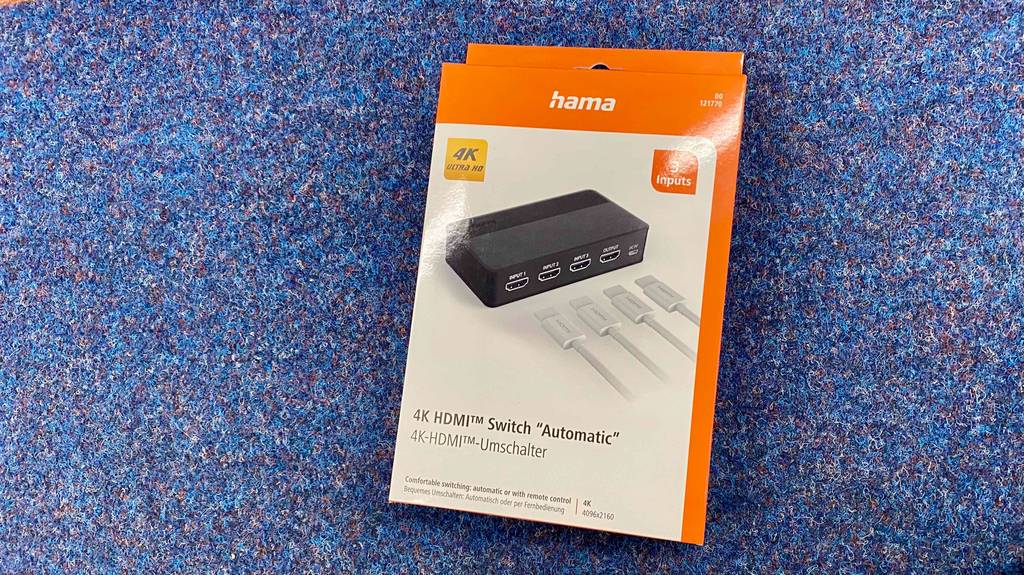 Ein verpackter HDMI Switch von Hama im Test liegt auf blauem Boden.