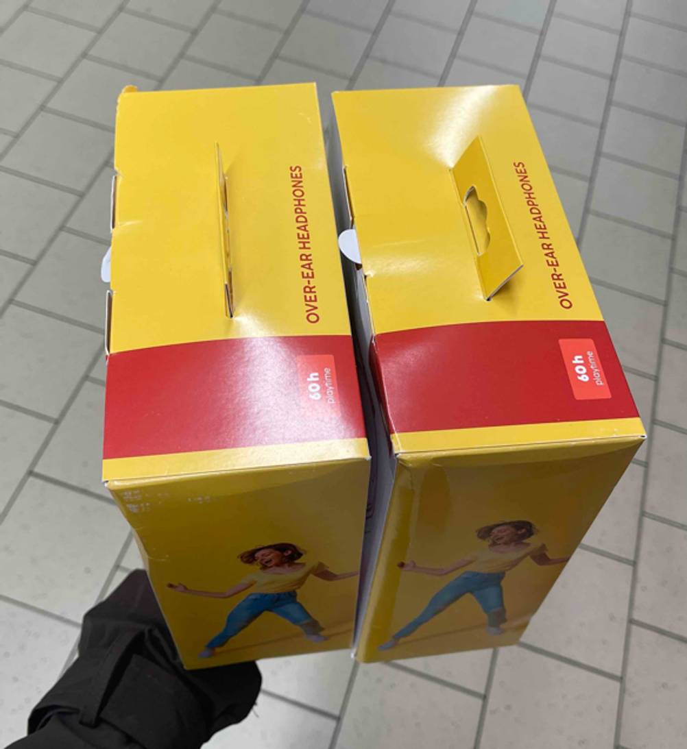 Eine Hand hält zwei Verpackungen der getesteten Hama-Kopfhörer „Spirit Calypso III“ mit 60 Stunden Batteriedauer über einem weißen Fliesenboden. 