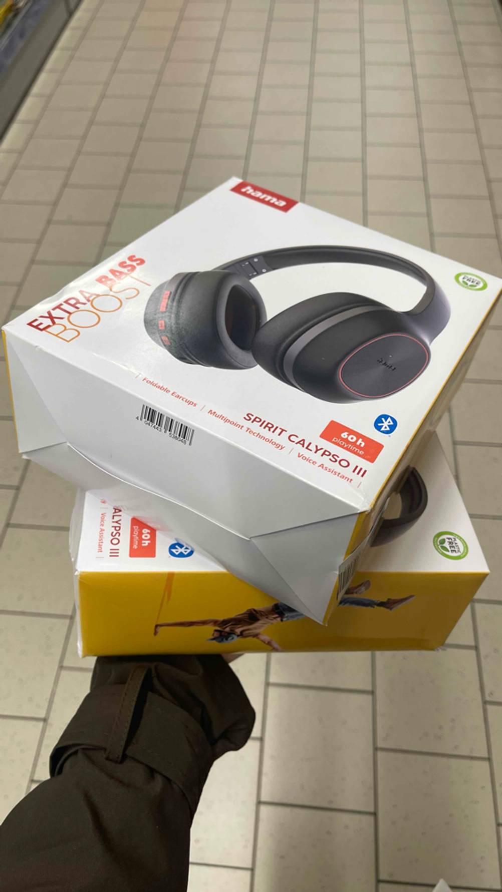 Eine Hand hält zwei Verpackungen der getesteten Hama-Kopfhörer „Spirit Calypso III“ über einem weißen Fliesenboden.