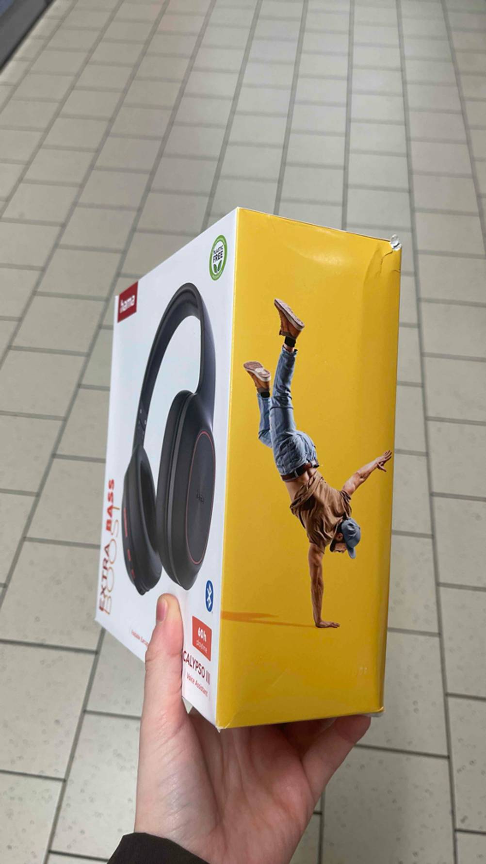 Eine Hand hält die gelb umrandete Verpackung des Hama-Kopfhörers im Test über einem weißen Fliesenboden. Es ist das Foto eines über Kopf stehenden jungen Mannes zu erkennen. 