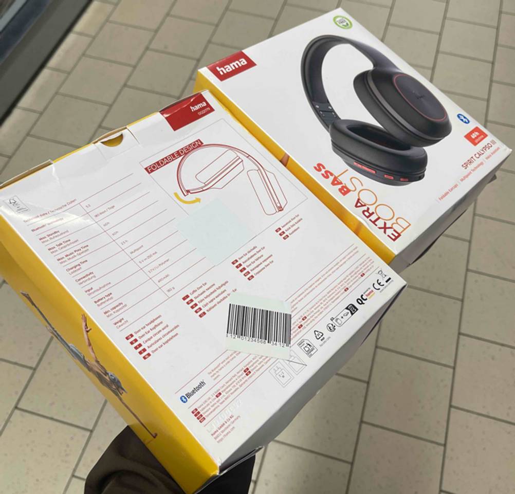 Eine Hand hält zwei nebeneinanderliegende Verpackungen der getesteten Hama-Kopfhörer „Spirit Calypso III“ über einem weißen Fliesenboden. Es sind Vorder- und Rückseite zu erkennen.