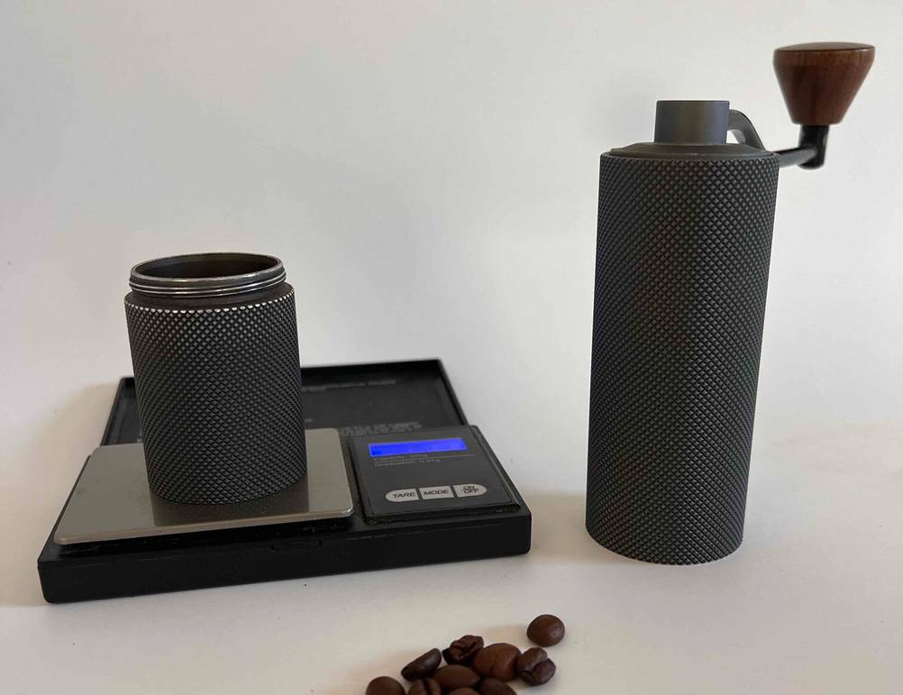 Auf einer Waage steht der untere Teil der getesteten Hand-Kaffeemühle, daneben der Teil mit der Kurbel, davor liegen einige Kaffebohnen.