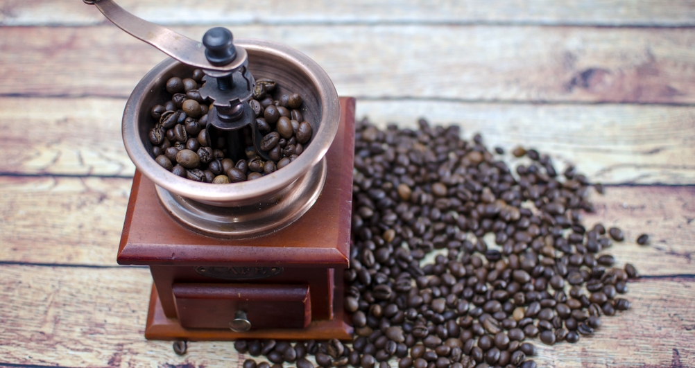 Eine Hand-Kaffeemühle steht auf einem Holztisch, daneben liegen Kaffeebohnen.