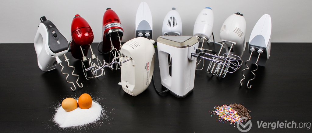 Handmixer-Test: Die getesteten Handmixer stehen nebeneinander auf einem schwarzen Tisch, davor Mehl, Eier und Schokostreusel.