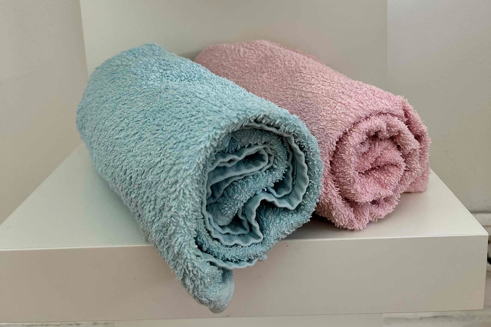 Handtücher im Test: Zwei zusammengerollte Handtücher in mint und rosa liegen schräg nebeneinander in weißem Regal.