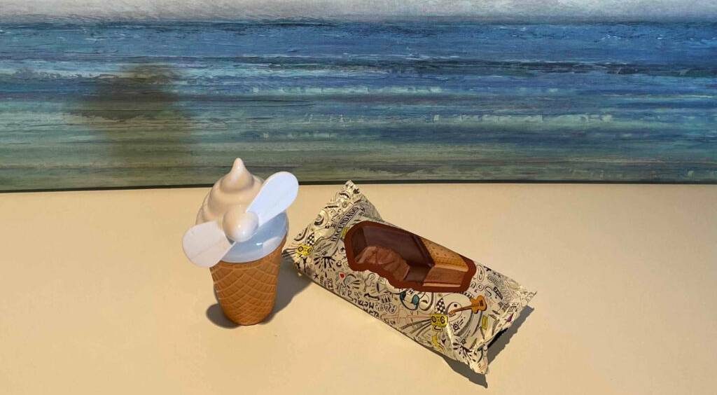 Vor einem Gemälde mit Meerlandschaft liegt ein getesteter Eishörnchen-Handventilator neben einer Eiswaffel in der Verpackung.