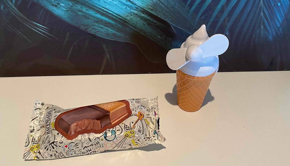 Handventilator in Form einer Eis-Waffel-Tüte steht neben einem Eiskeks vor einem Palmenbild.