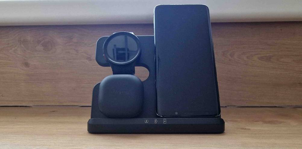 Schwarze Handy-Ladestation im Test befindet sich inklusive Smartwatch sowie In-Ear-Kopfhörer auf einem Holzuntergrund.