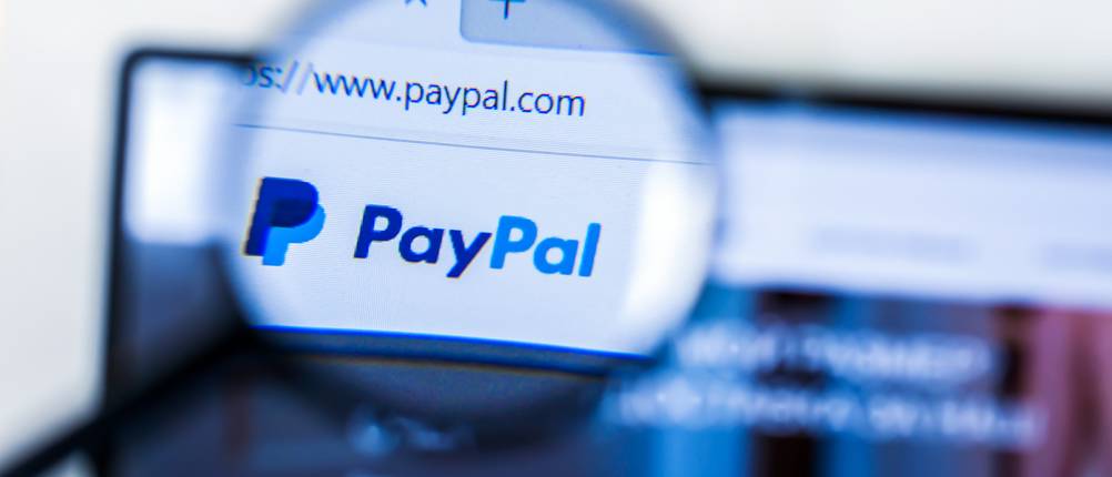 Durch eine Lupe ist das vergrößerte Logo von PayPal auf der Homepage zu sehen.