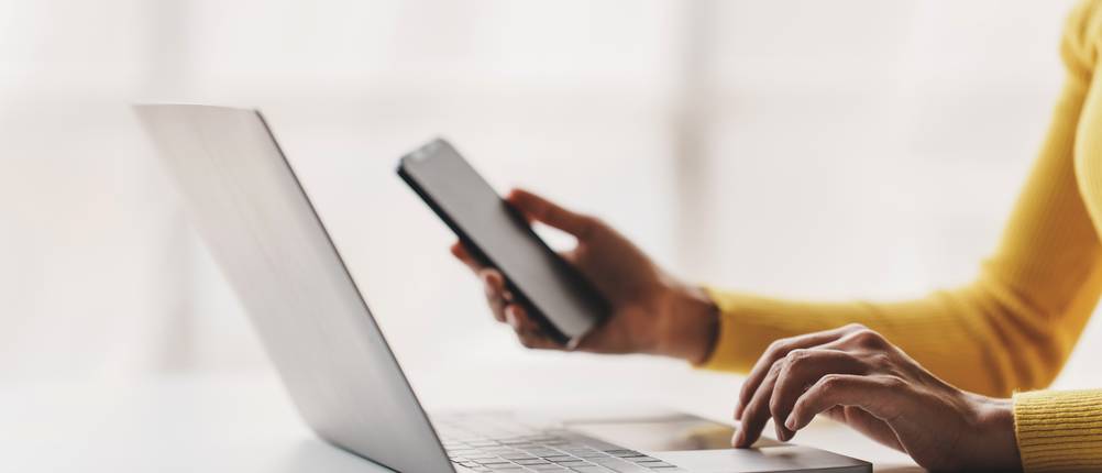 Eine Frau sitzt am Laptop und hält ihr Handy in der Hand.