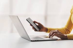 Anbieter zum Handy verkaufen im Test: Eine Frau sitzt am Laptop und hält ihr Handy in der Hand.