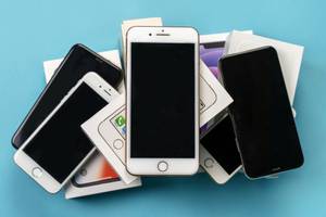 Handyversicherungen im Test: Mehrere alte iPhone-Modelle und die jeweiligen Verpackungen liegen auf einem türkisfarbenen Untergrund.