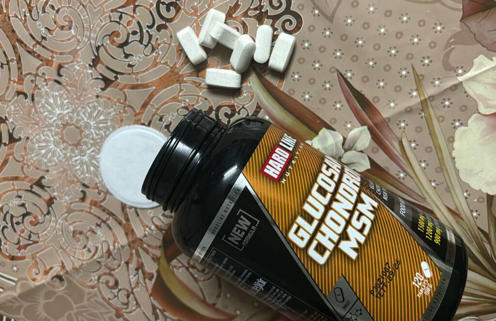 Getestetes Glucosamin in Tablettenform liegt neben einem schwarzen Flakon der Marke Hardline auf einer beige gemusterten Tischdecke.