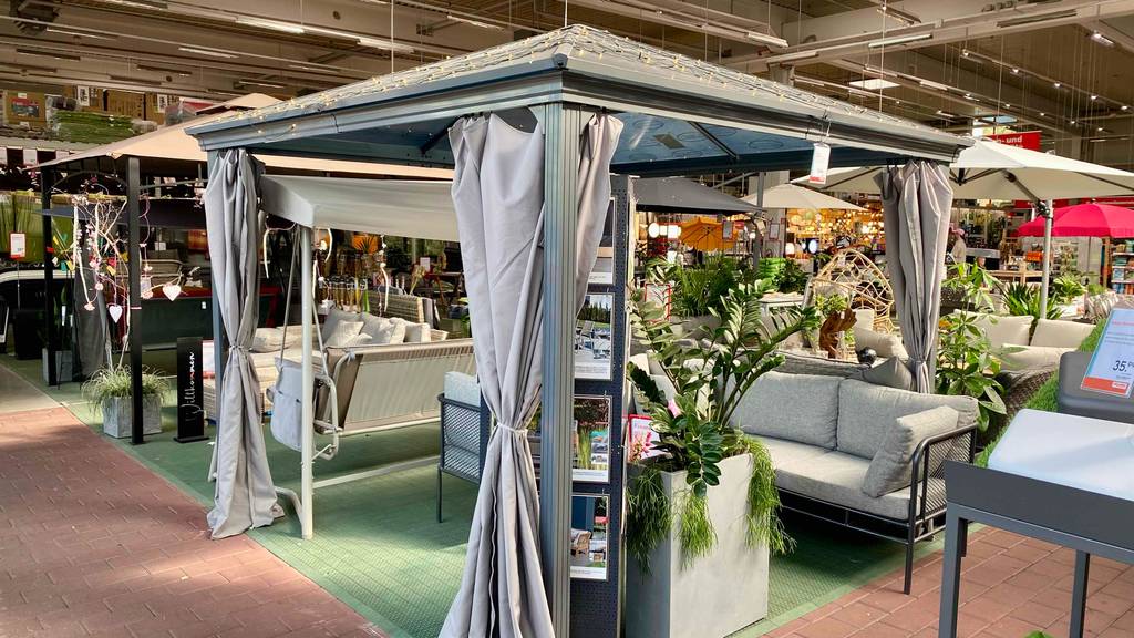Gesamtansicht übers Eck eines Metall-Pavillons im Test, der in einem Gartencenter aufgebaut und mit anthrazitfarbenen Vorhängen versehen ist.
