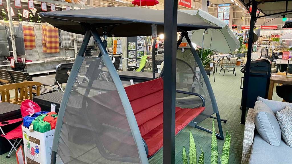 Hollywoodschaukel-Test: seitliche Sicht auf ein bedachtes Modell mit grünem Gestell und roter Polsterauflage, das zwischen Gartenmöbeln in einem Baumarkt steht.