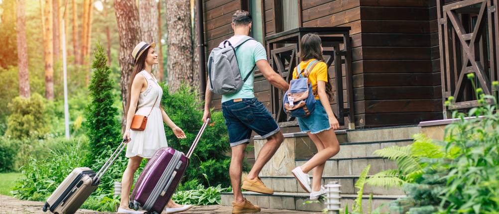 Familie zieht Koffer hinter sich her und steigt die Stufen zu einem Haus hoch.