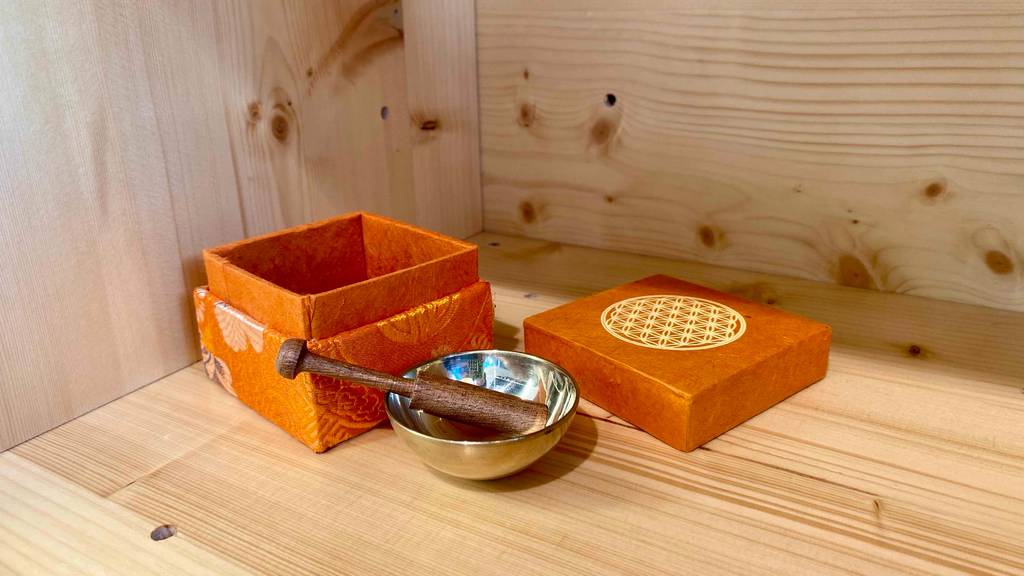 Im Test: Deckel und Unterteil einer Box liegen hinter der Klangschale mit Klöppel in einem Holzregal.