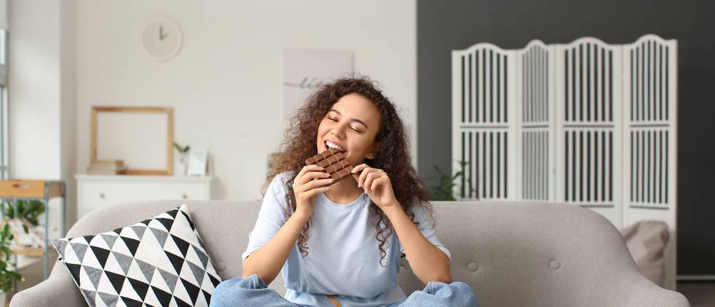 Eine junge Frau sitzt auf dem Sofa und beißt in eine Tafel Schokolade