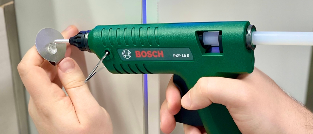 Die Bosch PKP 18 E Heißklebepistole wird auf eine kleine silberne Dekofläche mit schmalem Rand aufgetragen, gehalten von einer Hand.