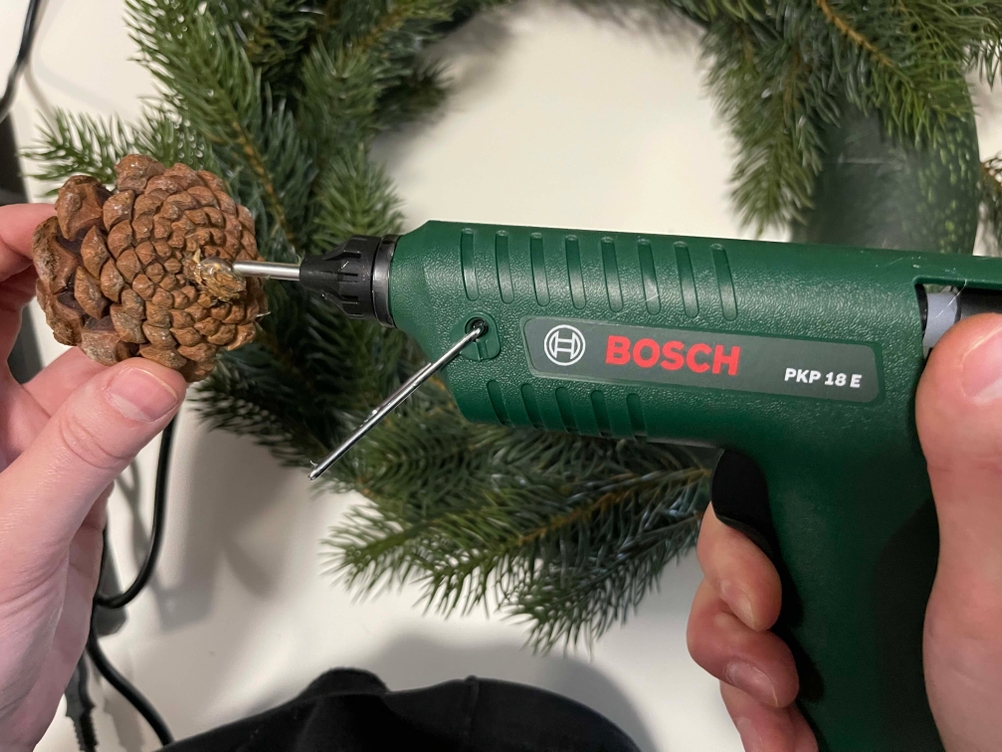 Bosch PKP 18 E Heißklebepistole klebt einen Tannenzapfen in einem DIY-Weihnachtsprojekt vor grünem Tannenzweig
