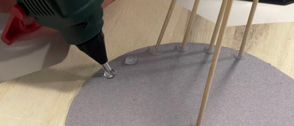 Parkside Heißklebepistole beim punktuellen Auftragen von Kleber auf eine graue Bastelplatte mit Holzstäben