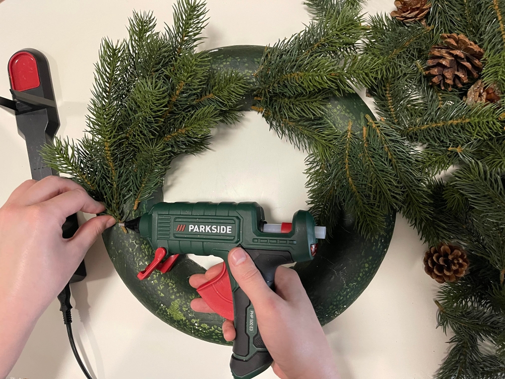 Parkside Heißklebepistole klebt Tannenzweig an grünen Deko-Kranz mit Zapfen und Kugeln