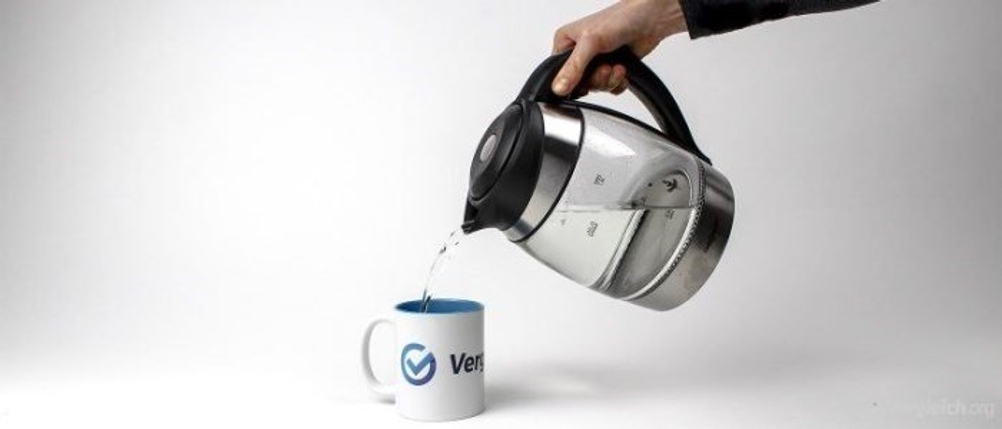 Eine Hand gießt mit einem Wasserkocher Wasser in eine Vergleich.org-Tasse.