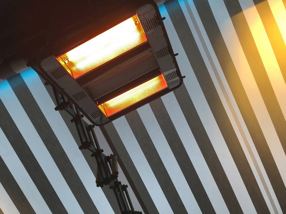 Heizpilz elektrisch im Test: Ein Infrarotstrahler in Funktion ist unter einer ausgefahrenen, grau-weiß gestreiften Markise zu sehen.
