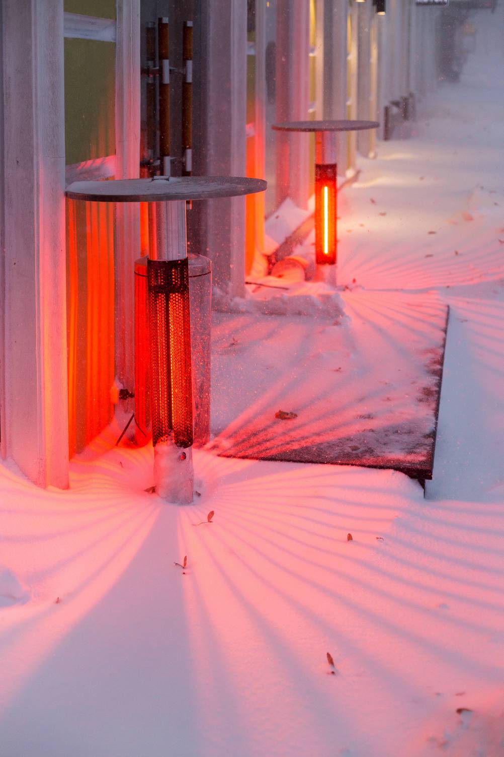 Heizpilz elektrisch im Test: Zwei elektrische Heizpilze mit Tisch stehen im Schnee vor einem Eingang und leuchten rötlich.