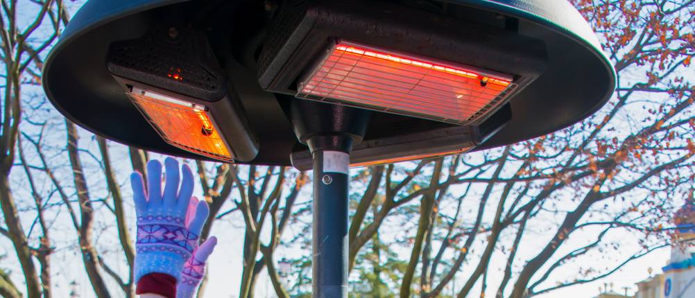 Heizpilz elektrisch im Test: Ein Heizpilz in Pilzform mit drei strahlenden Flächen. Unter einer sieht man behandschuhte Hände, die sich der Wärme entgegenstrecken.