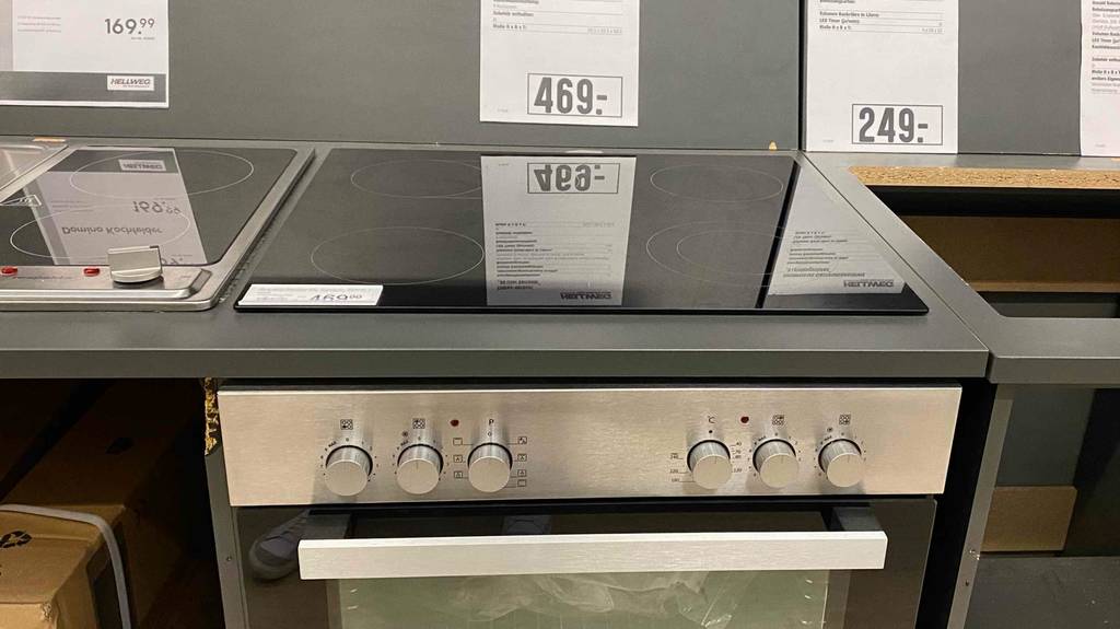 Ansicht von vorn auf die schwarze Kochplatte mit vier Kochzonen und den Backofen eines Hellweg Einbauherd-Sets im Test, der sich in einem Verkaufsbereich inklusive Preisschildern befindet.