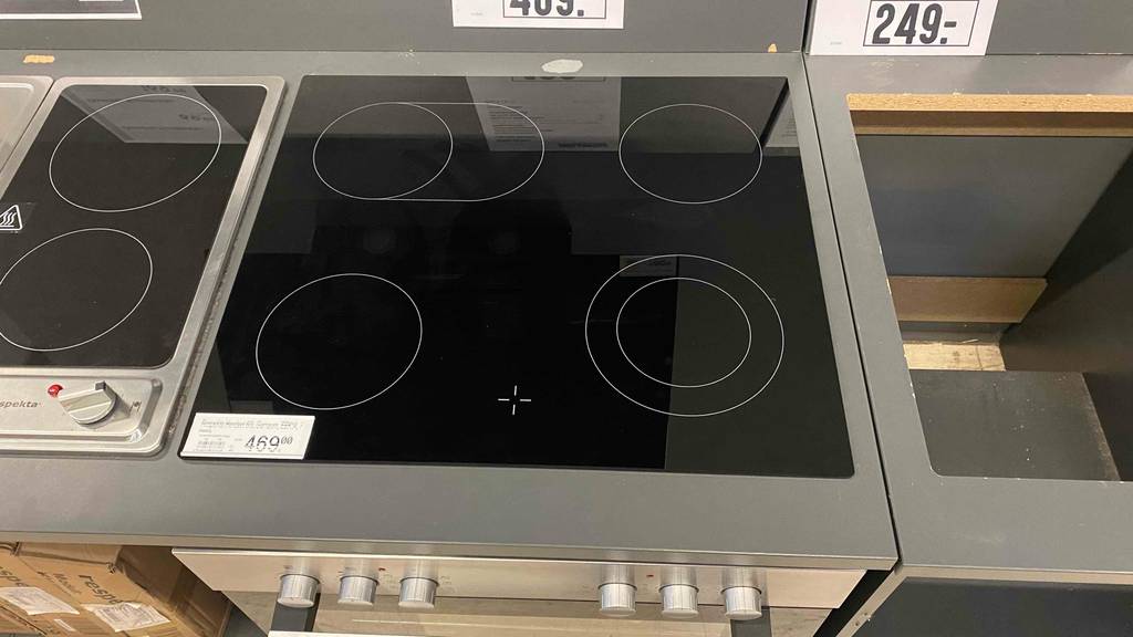 Ansicht von oben auf die schwarze Kochplatte inklusive vier Kochzonen eines Einbauherd-Sets im Test, der zwischen Kochplatten in einem Ausstellungsbereich steht.