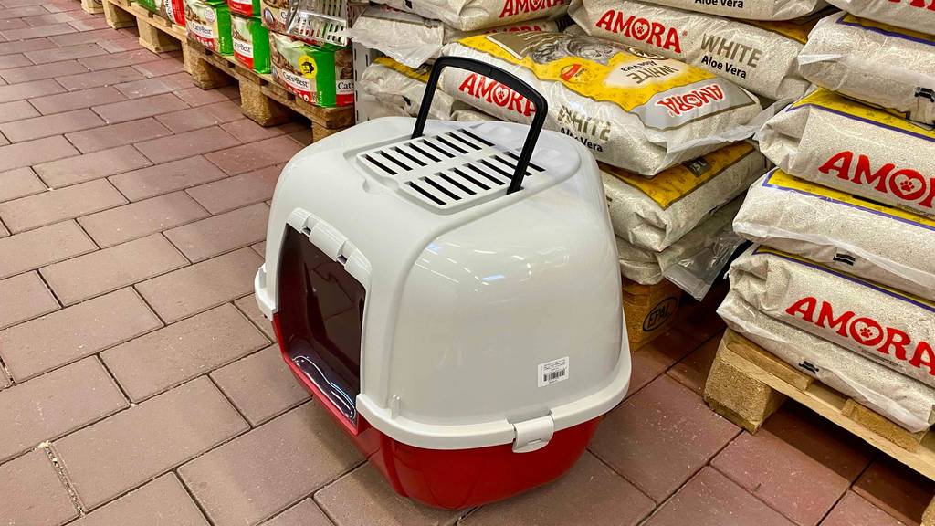 Ansicht von oben auf eine geschlossene Katzentoilette im Test, mit roter Auffangschale, weißem Deckel und transparenter Eingangsklappe sowie schwarzem Transportgriff, vor Katzenstreu-Verpackungen.
