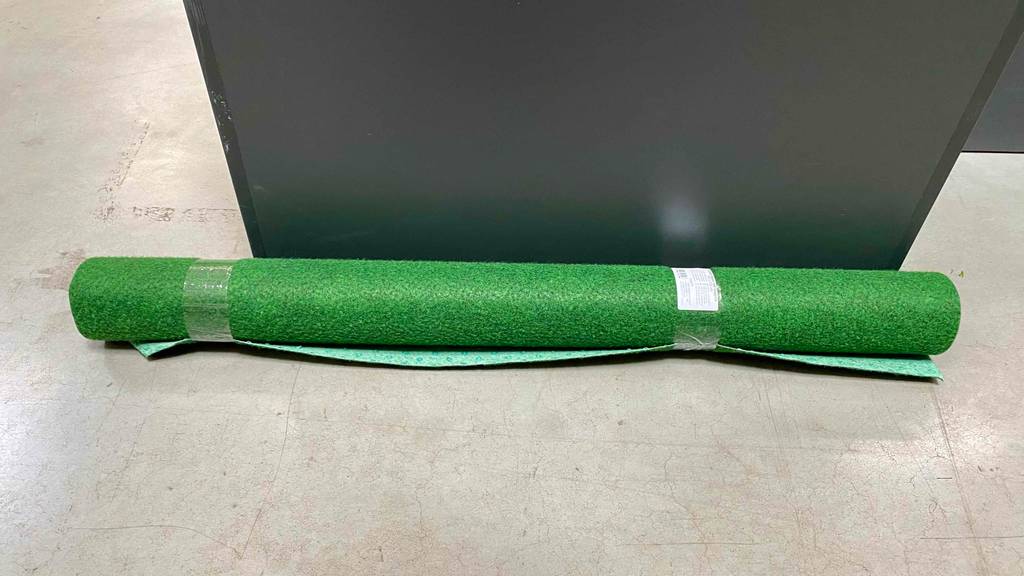 Eine Rolle grüner Kunstrasen im Test liegt auf einem Zementbelag vor einem schwarzen Behälter.