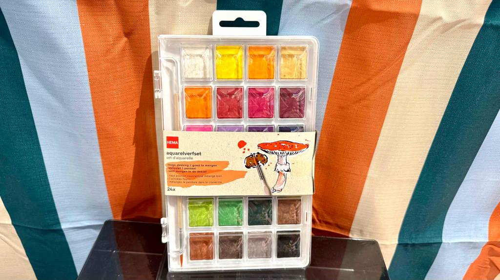 Vor einem farbig gestreiftem Gewebe steht eine Aufbewahrungsbox mit den getesteten Aquarellfarben.