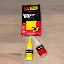 Das HG Power Glue Set mit beiden Fläschchen und Verpackung liegt geöffnet auf einem Holzboden. Die Produkte sind gut sichtbar platziert.