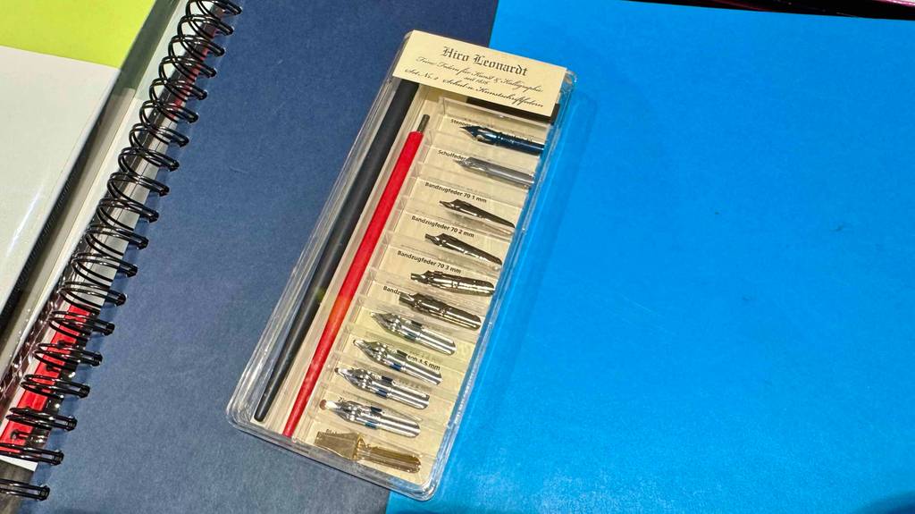 Getestete Kalligrafie-Stifte: Eine transparente Verpackung mit mehreren Wechselfedern und zwei Stiften liegt auf einem blauen Ringbuch.