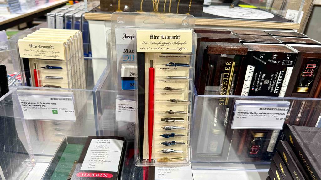 Im Test: Die Kalligrafie-Stifte in der transparenten Verpackung mit mehreren Wechselfedern lehnen an einer Box mit verschiedenen Arten von Kalligrafie-Stiften.