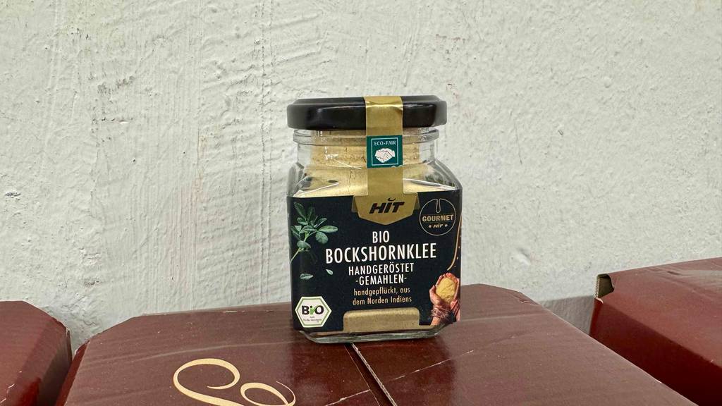 Eine Glasdose mit Bockshornklee im Test steht vor einer weißen Wand.
