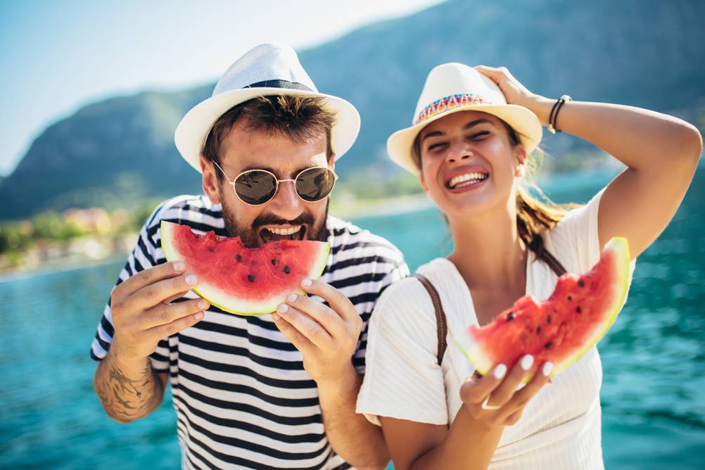 Eine Mann und eine Frau mit Sommerhüten essen Wassermelone vor einem See.