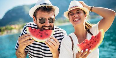 Eine Mann und eine Frau mit Sommerhüten essen Wassermelone vor einem See.