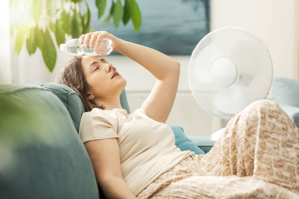 Eine Frau liegt erschöpft auf einer Couch. Sie hält sich zur Abkühlung eine kleine Wasserflasche an die Stirn. Die Augen sind geschlossen. Im Hintergrund läuft ein Ventilator