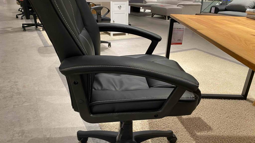 150-kg-Bürostühle-Test: Detailansicht von der Seite auf die Armlehnen eines schwarzen Modells, mit Lederbezug und weißen Ziernähten, das vor einem Holzschreibtisch auf beigefarbenem Teppichboden im Verkauf steht.