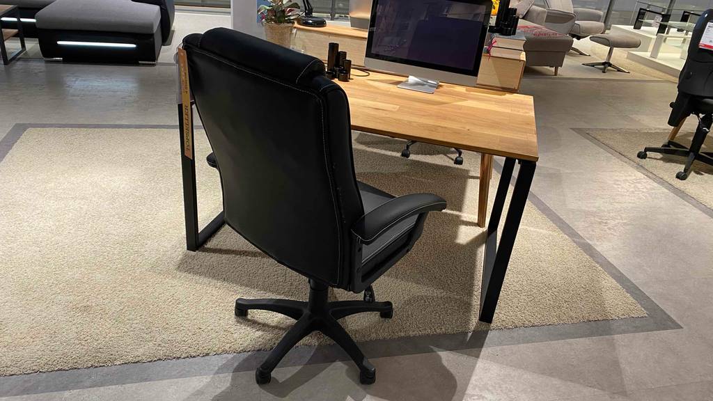 150-kg-Bürostühle im Test: Ansicht von hinten auf die hohe Rückenlehne von einem schwarzen Modell auf Rollen vor einem Schreibtisch mit einer Tischlatte aus Holz in einem Verkaufsbereich.