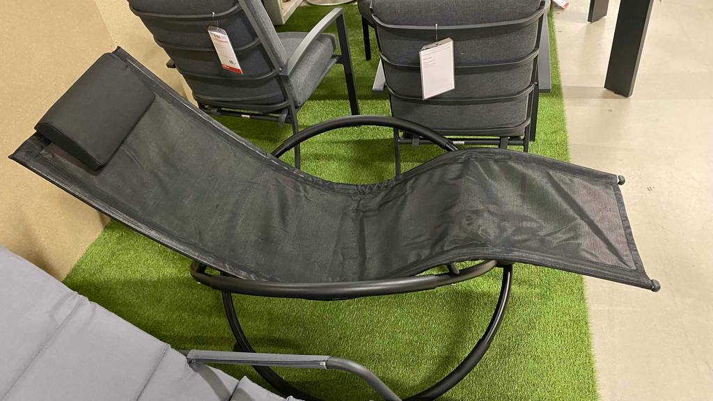 Schaukelliegen-Test: eine schwarze Schaukelliege mit schwarzem Gestell und wasserabweisendem Bezug steht auf einem Kunstrasen im Verkaufsbereich neben Outdoor-Sesseln.