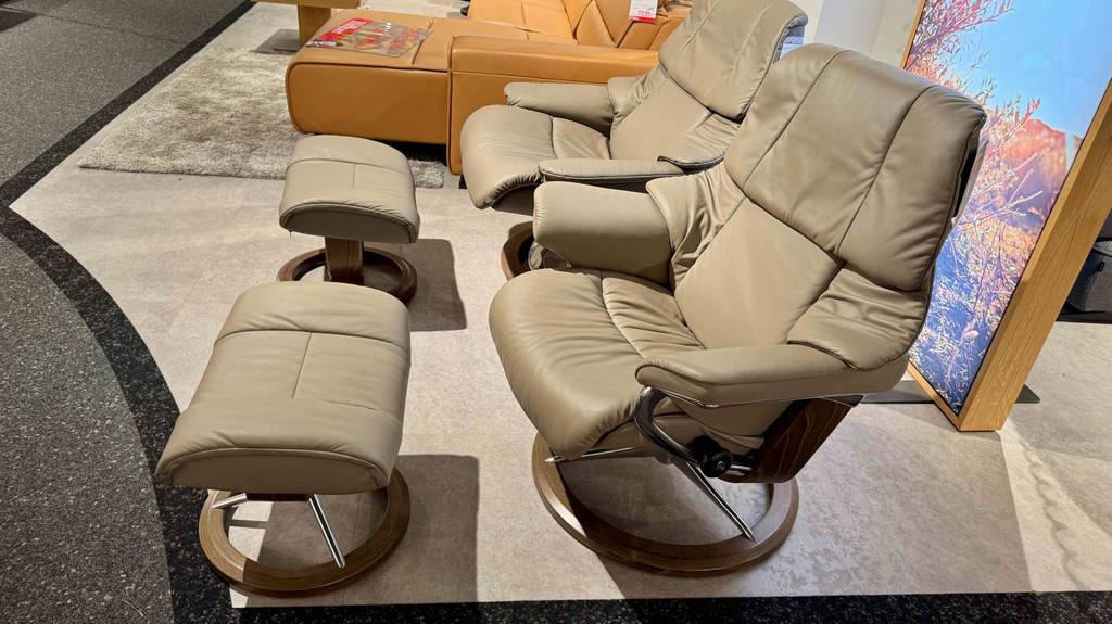 Ein getesteter Relaxsessel mit Hocker in beigefarbener Lederausführung, von der Seite betrachtet, steht neben einem Sofa.