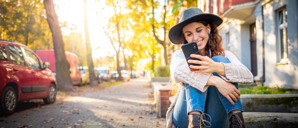 Junge Frau mit Hut und In-Ear-Kopfhörern sitzt im Freien auf einer Stufe und hält ein Smartphone
