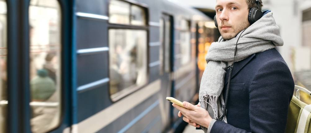 Mann mit Kopfhörern steht mit einem Smartphone in der Hand an einem U-Bahnsteig